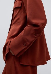 Rostfärgad skjorta med texturerad yta. Har en ficka och knäppning vid ärmsluten. Designen är avslappnad med en något oversized passform.