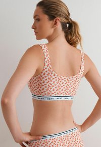 Bralet s cvjetnim printom u bijeloj boji s narančastim cvjetovima. Ima široku crnu elastičnu traku s logom "NEXT". Glatka tekstura, izrez u obliku školjke.
