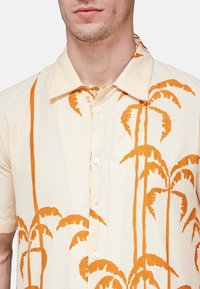 Kratka majica s kratkimi rokavi iz svetle bež tkanine s potiskom oranžnih palm, klasičen ovratnik in gumbasto zapiranje spredaj.