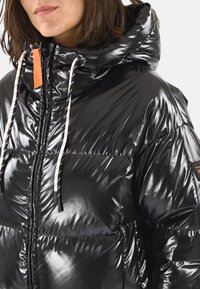 Doudoune noire avec une texture matelassée brillante. Dispose d'un col montant, d'une capuche avec cordon de serrage et d'une tirette de fermeture éclair orange contrastante.