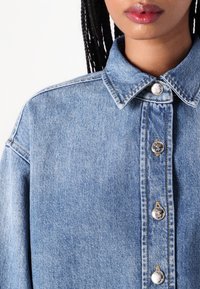 Camicia in denim azzurro chiaro con colletto a punta, chiusura frontale con bottoni e dettagli metallici, e tessuto testurizzato.