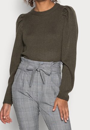 Femme portant un pull en tricot olive foncé à épaules bouffantes et un pantalon gris à carreaux taille haute, noué à la taille avec une ceinture en forme de nœud.