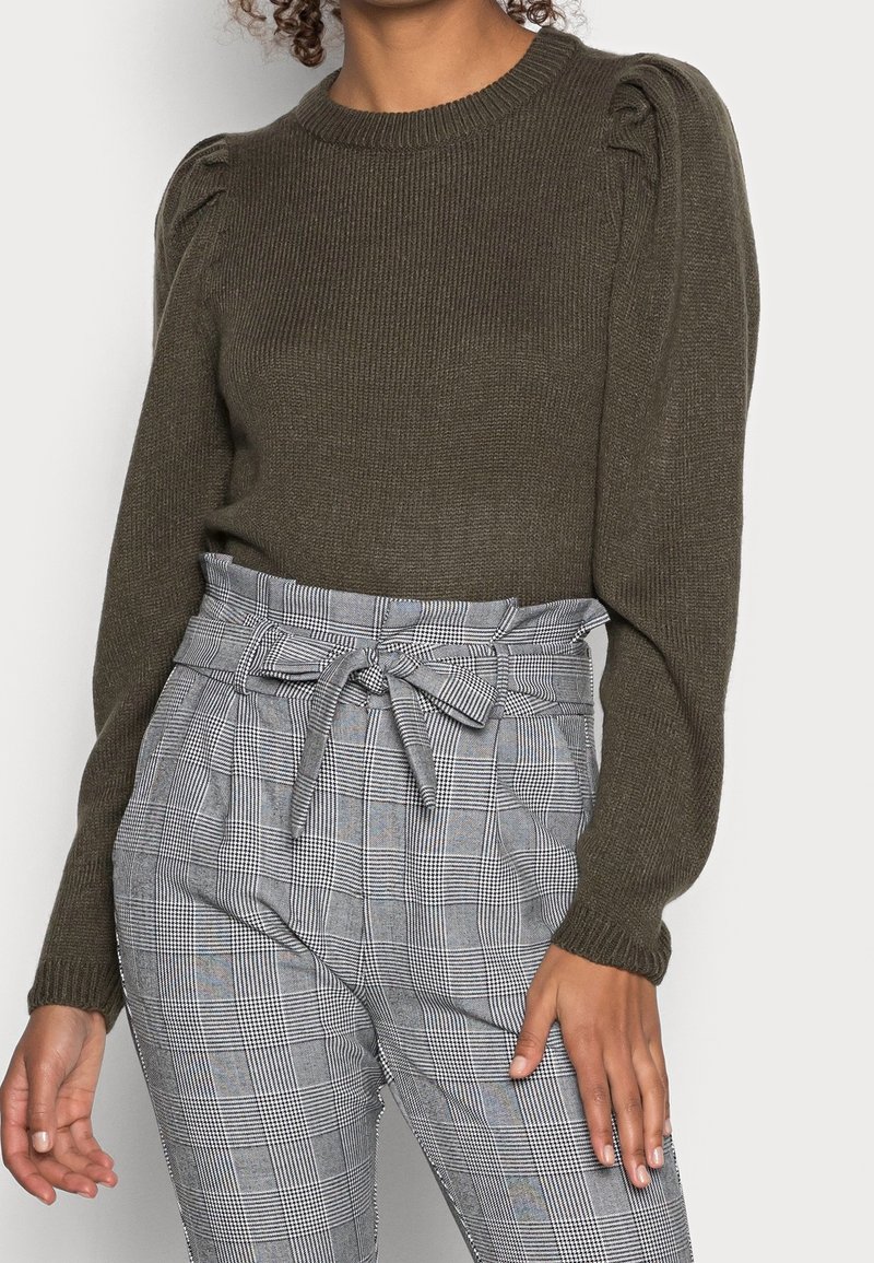 Femme portant un pull en tricot olive foncé à épaules bouffantes et un pantalon gris à carreaux taille haute, noué à la taille avec une ceinture en forme de nœud.