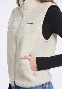 Cremfarbener Fleece-Weste mit hohem Kragen, Reißverschluss vorne und Seitentaschen. Mit einem Logo-Patch auf der Brust und einer strukturierten Oberfläche.