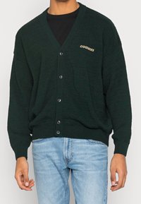 Mann in einer dunkelgrünen, knöpfbaren Strickjacke mit dem Text "GOODBOIS" auf der Brust und hellblauen Jeans, der vor einem weißen Hintergrund steht.