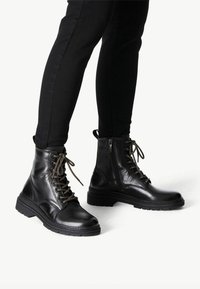 Des bottes de combat en cuir noir présentent un laçage à l'avant, une fermeture éclair sur le côté, un bout rond, une semelle texturée et des lacets assortis, conçues pour la durabilité et le confort.