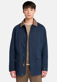 Chaqueta azul marino con un cuello de pana marrón, botones de presión, dos bolsillos frontales y una tela texturizada. Presenta un cierre con cremallera y costuras en contraste.