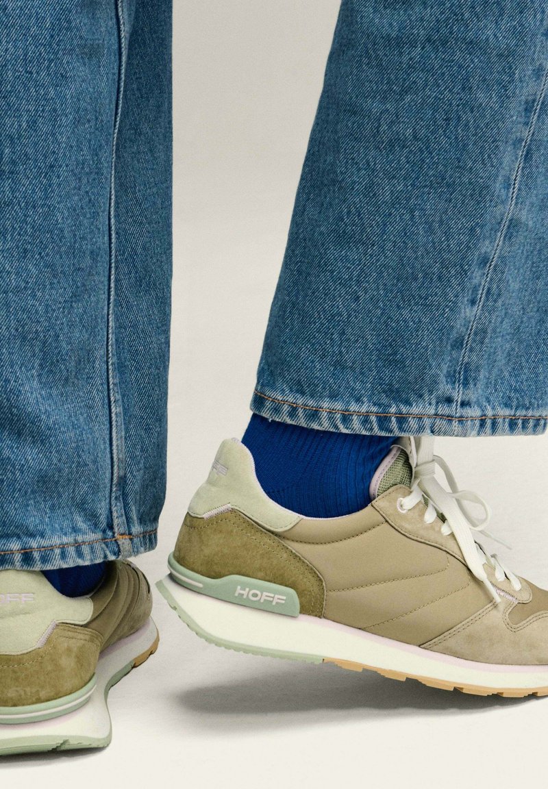 Zapatillas beige y verde oliva con cordones blancos, detalles de ante texturizado y suelas verde claro, combinadas con calcetines azules y pantalones de mezclilla.