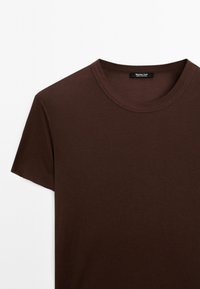 Hnědé bavlněné tričko s kruhovým výstřihem a krátkými rukávy, které má hladkou texturu a jednoduchý design. Nálepka umístěná na výstřihu.