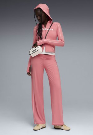 Woman wearing a pink hooded zip-up jacket and matching wide-leg pants, holding a small white Puma handbag, beige sneakers on gray background.