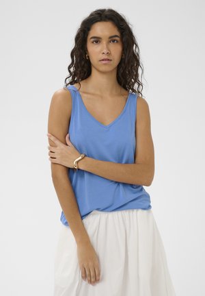 Vrouw met krullend donker haar, gekleed in een blauwe mouwloze top en witte rok, poserend met één arm gekruist en een gouden armband om haar pols.