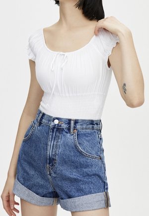 Kvinde iført en hvid kortærmet, rynket top og højtaljede, opsmøgede denimshorts med en tatovering på venstre underarm.