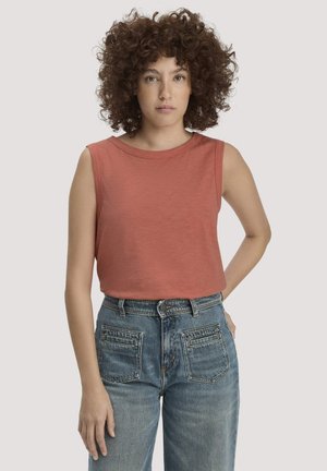 Donna con capelli ricci che indossa una canotta color ruggine e jeans blu a vita alta, in piedi con una mano sui fianchi.