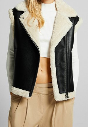 Vrouw met een mouwloos zwart leren vest met witte shearling voering over een wit geribd cropped truitje en beige geplooide broek.