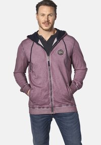 Purple zip-up hoodie with a navy interior hood and logo. Features front pockets, cuffed sleeves, and a relaxed fit. Worn over a black shirt.