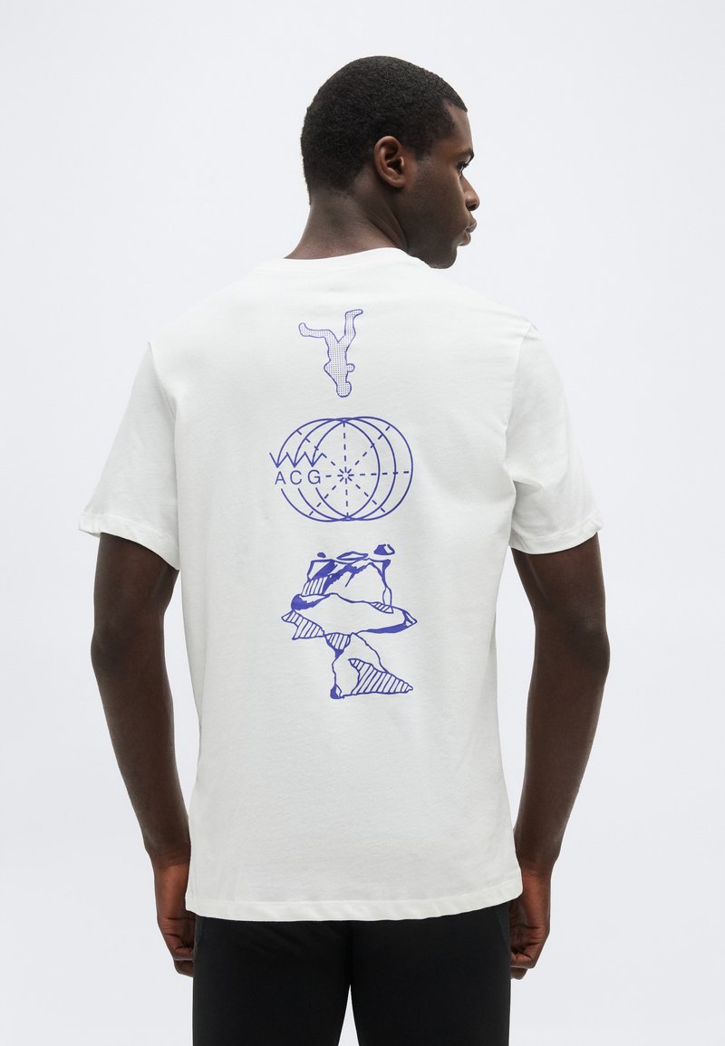 Άνδρας που φοράει λευκή φανέλα με μπλε αφηρημένα σχέδια: αναποδογυρισμένο πρόσωπο, γήινη σφαίρα με το κείμενο "ACG" και ακανόνιστα σχήματα στην πλάτη.