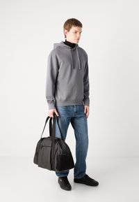 Grijze hoodie met trekkoorden, blauwe denim jeans, zwarte sneakers en een zwarte stoffen sporttas met handvatten bovenaan. Eenvoudige, casual look.