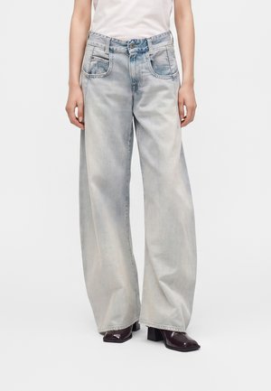 Person iført lyse, vidde jeans, hvid top og mørke støvler med firkantet tå og hæl, stående mod en ensfarvet baggrund.