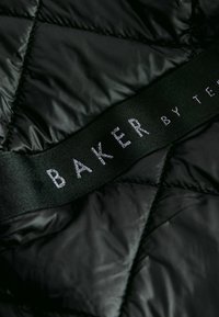 Crni kvadratični materijal s sjajnom teksturom sadrži istaknutu crnu oznaku u kojoj je urezano "BAKER BY T." srebrnim slovima.