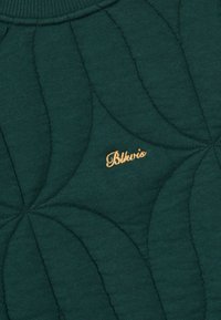 A zöld steppelt anyag kiemelkedő levélmintákkal és aranyhímzett logóval rendelkezik, ami textúrát és vizuális érdekességet ad a dizájnnak.