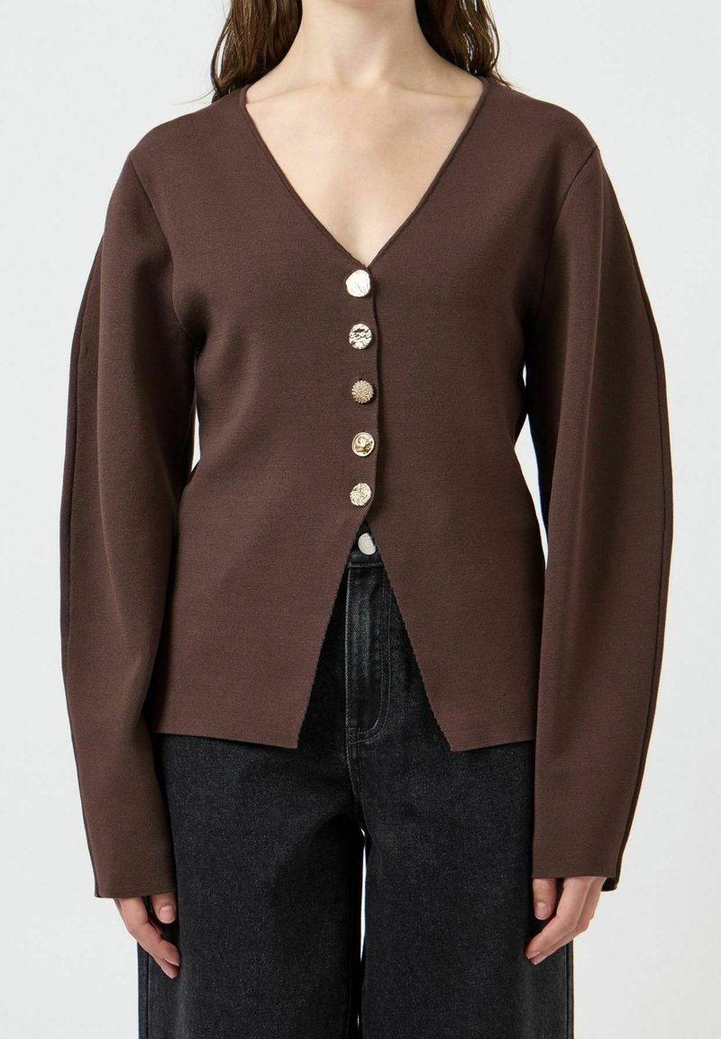 Femme portant un cardigan marron à col en V avec cinq boutons uniques, associé à un jean noir taille haute, présenté de la poitrine aux hanches.