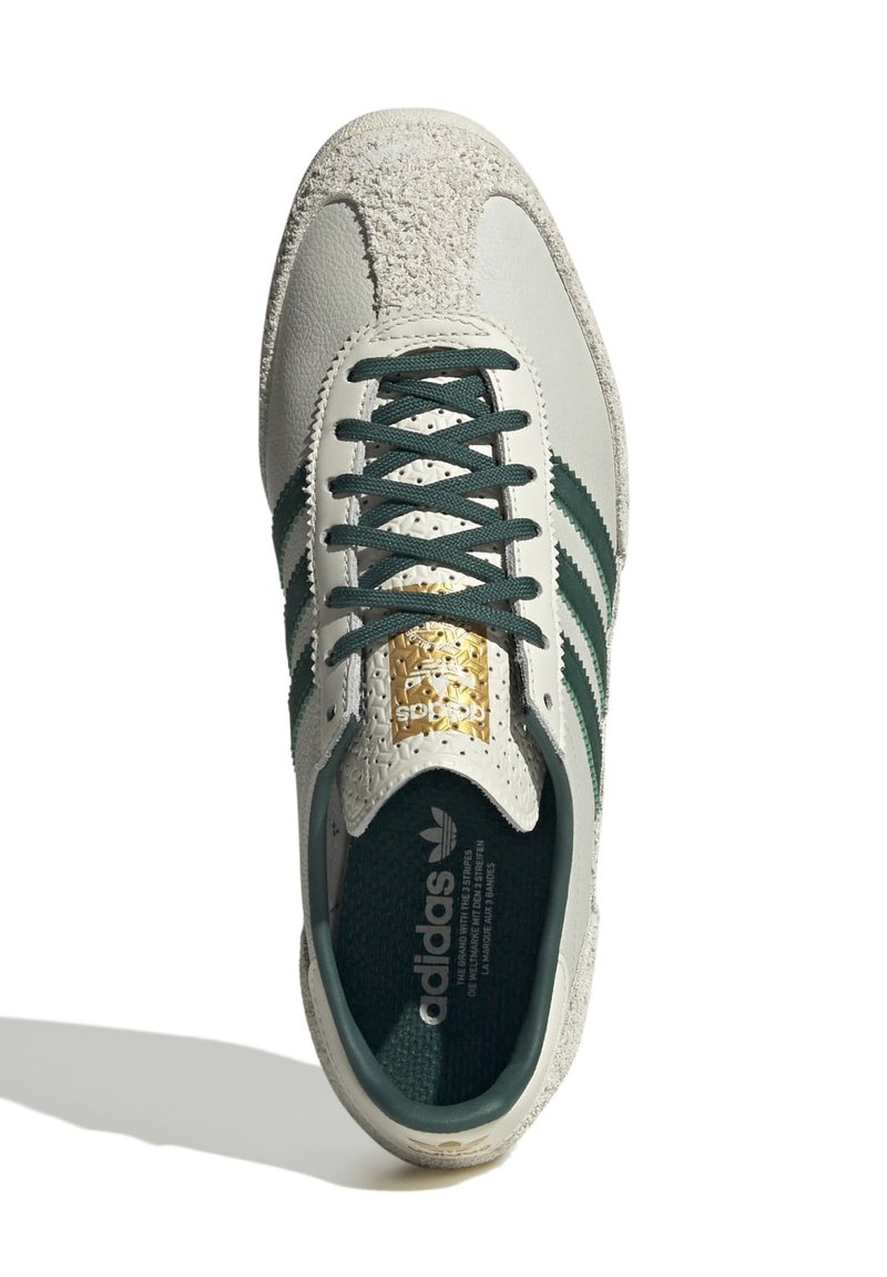 Sneaker en cuir gris clair avec des rayures vertes, capuchon en suède texturé, accents dorés sur la languette et semelle intérieure à motifs. Design à lacets.