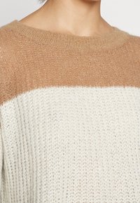 Pull en tricot avec un design en color-block, la partie supérieure est beige et la partie inférieure est crémeuse. Texture douce avec un col rond et une coupe ample.