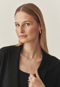 Złote kolczyki o rzeźbiarskim wzorze, noszone przez osobę w czarnym swetrze. Widoczne są gładka skóra, proste włosy i minimalny makijaż.