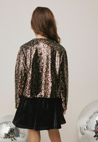 Paillettenjacket met een zwart en gouden patroon, getextureerd met glinsterende pailletten, gecombineerd met een zwarte fluwelen rok. Zilveren discoballen op de achtergrond.