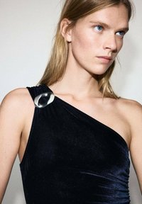 Vestido de terciopelo negro con un tirante en el hombro, adornado con un acento circular plateado, textura suave y silueta ajustada.