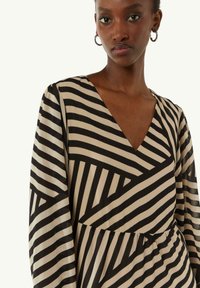 Schwarz- und cremefarben gestreiftes Kleid mit V-Ausschnitt und langen Ärmeln. Verfügt über unterschiedliche diagonale Linienmuster und eine geraffte Taille.