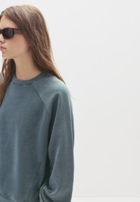 Teal sweatshirt med raglanærmer, lavet af blødt stof, har en ribbet halsudskæring. Bæres med mørke rektangulære solbriller.