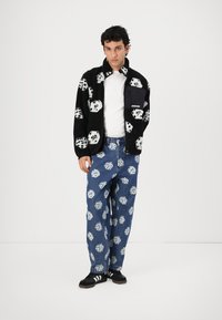 Black fleece jacket with white dice pattern, paired with blue denim pants featuring similar dice print. Black sneakers complete the outfit.