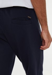 Pantalones de chándal en color navy que presentan una tela texturizada, cremallera lateral y un pequeño bolsillo trasero. El diseño incluye un sutil detalle de logo.