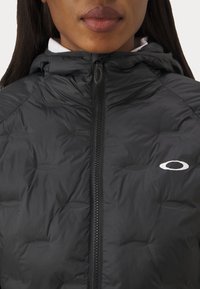 Schwarze Pufferjacke mit Reißverschluss vorne, Kapuze, gestepptem Muster und kleinem weißen Logodetail auf der linken Brust.