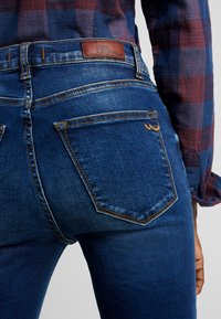 Vaqueros de mezclilla azul con un parche de cuero marrón del logo LTB. Presenta bolsillos traseros clásicos y detalles de costura sutiles. Diseño ajustado.