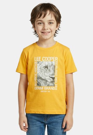 Jeune garçon souriant, portant un t-shirt jaune avec un lion graphique et un texte blanc indiquant "LEE COOPER DENIM BRAND" ainsi qu'un jean bleu.