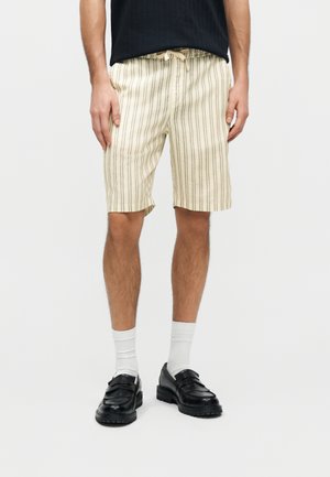 Muž nosiaci krémové šortky s tenkými vertikálnymi pruhmi, biele ponožky, čierne kožené mokasíny a čierne tielko bez rukávov.