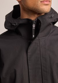 Chaqueta negra impermeable con un alto cuello, capucha ajustable y cremallera central con un tirador texturizado, que presenta un diseño minimalista.