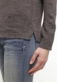 Brun stickad tröja med långa ärmar; har en sid Spricka och ribbad nederkant. Kombinerad med blekta blåjeans. Texturen verkar mjuk.