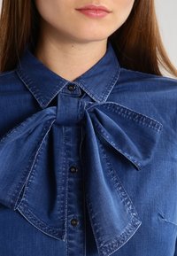 Jeansskjorta i mörkblått med en stor fluga-krage, knapplist fram och synliga sömnadsdetaljer, vilket ger en texturerad look.