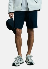 Marinefarbene Shorts kombiniert mit einer weißen Zip-Jacke. Das Modell hält eine schwarze Mütze. Die Schuhe haben silberne Akzente und strukturierte Sohlen.