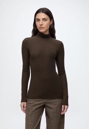 Vrouw met lang donker haar draagt een aansluitende bruine coltrui en losse bruine geruite broek, staand tegen een effen witte achtergrond.