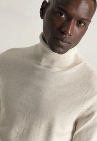 Primer plano de un hombre que lleva un suéter de cuello alto beige claro, mirando directamente a la cámara con una expresión neutral.