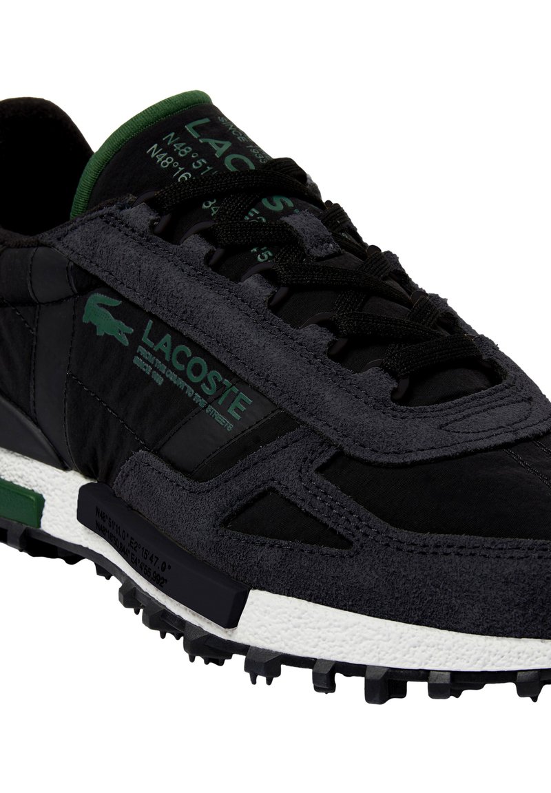Zapatilla Lacoste negra y gris oscuro con logo y texto en verde, cordones negros, suela blanca texturizada y dibujo de la suela resistente.