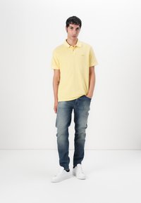 Gul polo skjorte med logo, kortermet og myk tekstur, kombinert med medium-wash denimjeans og hvite sneakers. Enkelt, casual utseende.