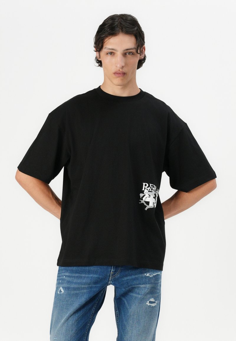 Camiseta negra de gran tamaño hecha de algodón con cuello redondo. Presenta un logo gráfico blanco impreso en el lado inferior izquierdo. Combinada con unos vaqueros azules desgastados.