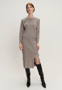 Vrouw in taupe gebreide trui en bijpassende rok met zijknopen en split, gecombineerd met zwarte kniehoge laarzen, staande voor een effen achtergrond.
