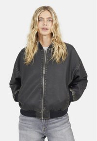 Giacca bomber nera con finitura lucida, polsini e orlo a costine, chiusura con zip e vestibilità comoda. Indossata sopra una maglietta grigia e abbinata a jeans.