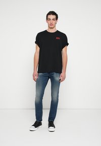 Czarny T-shirt z krótkim rękawem z podwiniętymi rękawami, z czerwonym logo "DIESEL", zestawiony z dopasowanymi niebieskimi jeansami i czarnymi tenisówkami.